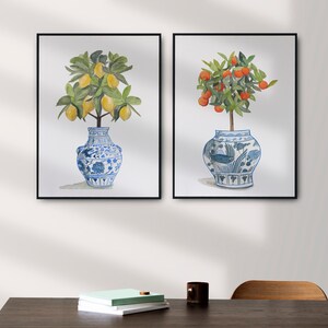 May include: Two framed watercolour paintings of potted citrus trees. The trees are in blue and white porcelain vases with intricate designs. The lemon tree has yellow lemons and the orange tree has orange fruit.
