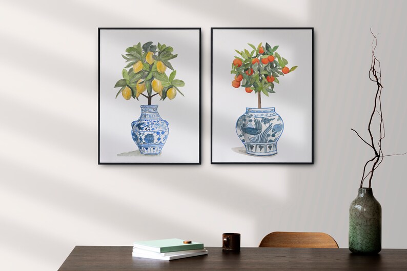 May include: Two framed watercolour paintings of potted citrus trees. The trees are in blue and white porcelain vases with intricate designs. The lemon tree has yellow lemons and the orange tree has orange fruit.