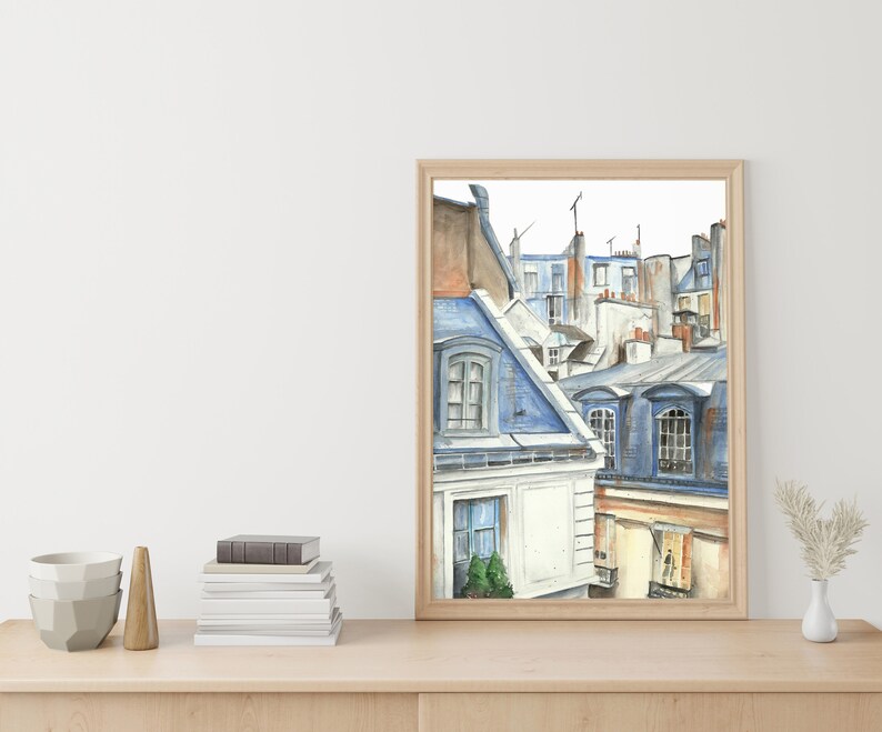 Puede incluir: Pintura acuarela de un paisaje urbano parisino con techos y ventanas azules y grises. La pintura est&aacute; enmarcada en un marco de madera clara y est&aacute; colgada en una pared blanca.