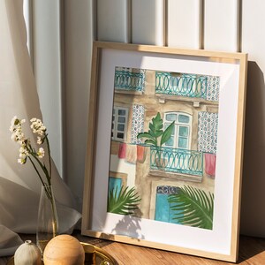 May include: A framed watercolour painting of a building with a balcony and a potted plant. The building is painted in shades of brown, blue, and white. The balcony has a teal railing and is decorated with colourful laundry hanging from it. The plant is a green palm tree. The painting is framed in a light wood frame.