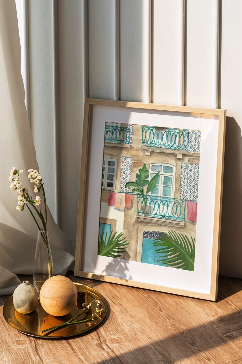 May include: A framed watercolour painting of a building with a balcony and a potted plant. The building is painted in shades of brown, blue, and white. The balcony has a teal railing and is decorated with colourful laundry hanging from it. The plant is a green palm tree. The painting is framed in a light wood frame.