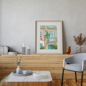 May include: A watercolour painting of a building with a balcony and a window. The building is painted in shades of beige and brown, with blue accents. The balcony has a green plant in a pot and a blue railing. The window is open and has a white frame. The painting is framed in a light brown frame.