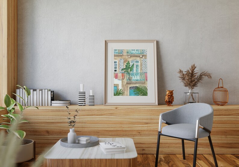 May include: A watercolour painting of a building with a balcony and a window. The building is painted in shades of beige and brown, with blue accents. The balcony has a green plant in a pot and a blue railing. The window is open and has a white frame. The painting is framed in a light brown frame.
