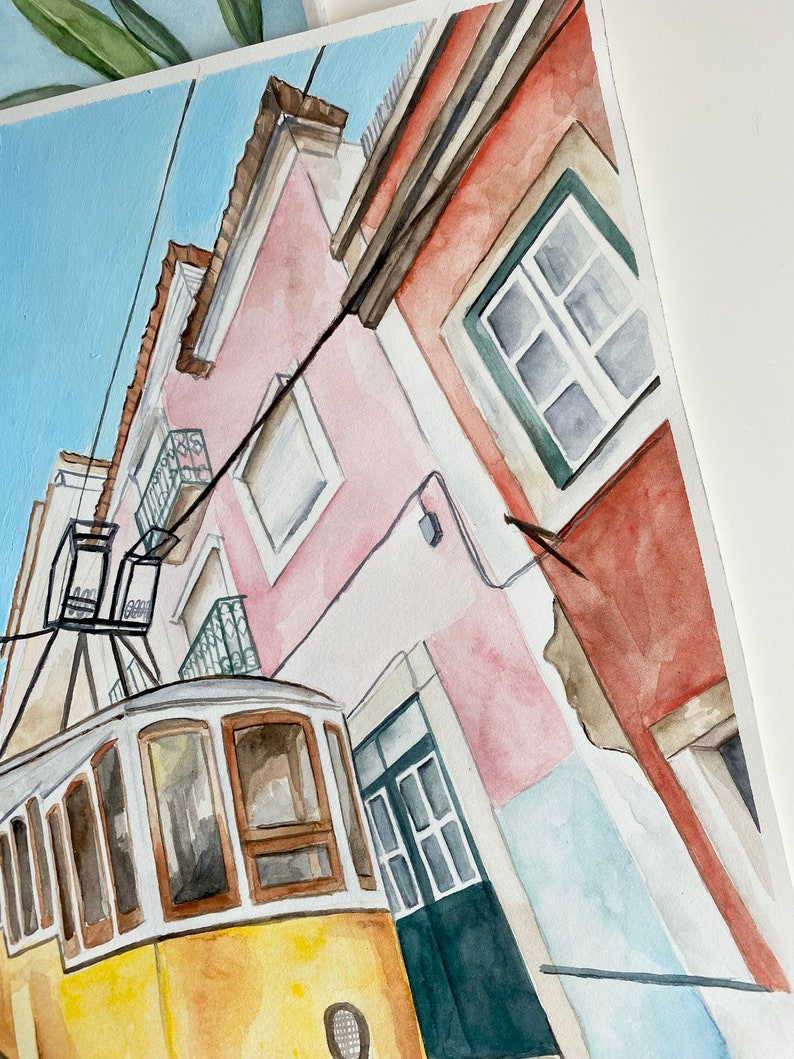May include: Watercolor painting of a yellow tram traveling up a narrow street lined with pink and red buildings. The buildings have windows and doors, and the tram has a white roof and brown trim.