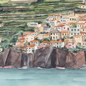 May include: Watercolor painting of a small village nestled on a cliff overlooking a body of water. The village is made up of colorful buildings with red tile roofs. The cliff is a dark brown color and the water is a light blue.