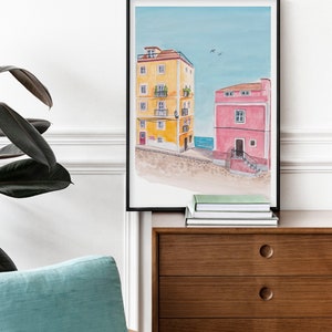 May include: A framed watercolor painting of two colorful buildings with a blue sky and sea in the background. The building on the left is yellow with white trim and the building on the right is pink with white trim.