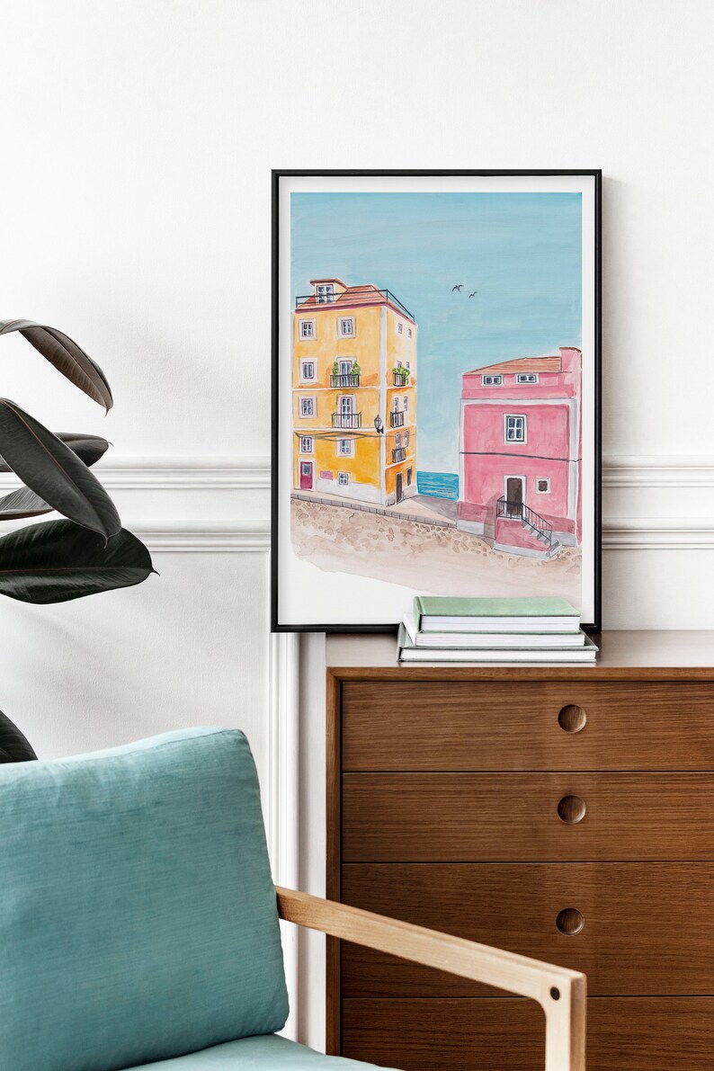 May include: A framed watercolor painting of two colorful buildings with a blue sky and sea in the background. The building on the left is yellow with white trim and the building on the right is pink with white trim.
