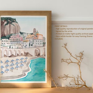 May include: Watercolor painting of a colorful Italian village on a cliff overlooking a beach with blue and white striped umbrellas. The painting has a white border and is framed in a light wood frame.