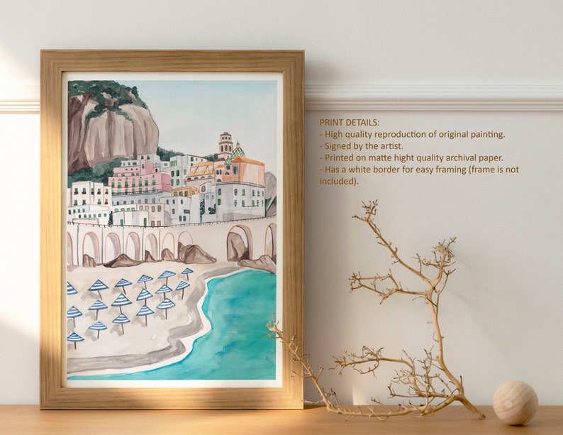 May include: Watercolor painting of a colorful Italian village on a cliff overlooking a beach with blue and white striped umbrellas. The painting has a white border and is framed in a light wood frame.