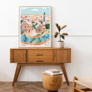May include: A framed illustration of a colorful cityscape with a mosaic tiled fountain in the foreground. The illustration is hanging above a wooden cabinet with two drawers. A potted plant sits on top of the cabinet.