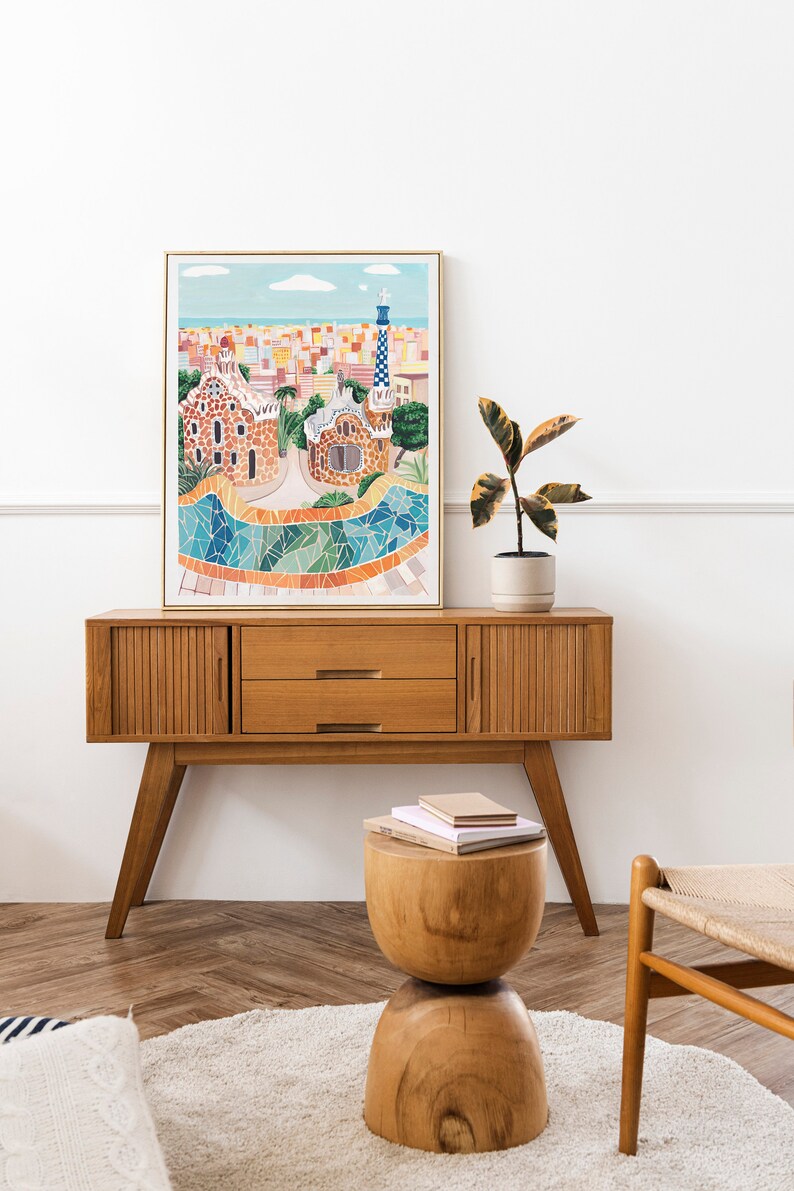 May include: A framed illustration of a colorful cityscape with a mosaic tiled fountain in the foreground. The illustration is hanging above a wooden cabinet with two drawers. A potted plant sits on top of the cabinet.