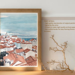 May include: Watercolor painting of a cityscape with red-tiled roofs and a blue sky. The painting is framed in a light wood frame and has a white border.