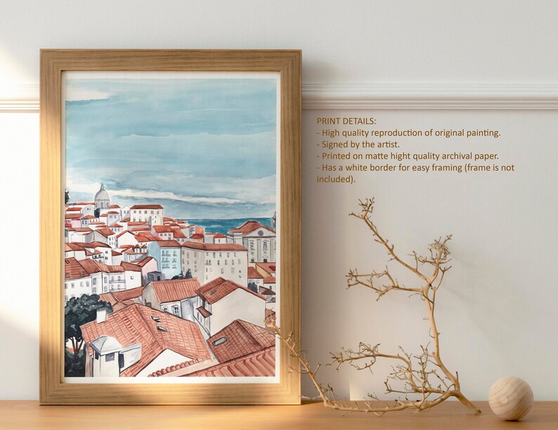 May include: Watercolor painting of a cityscape with red-tiled roofs and a blue sky. The painting is framed in a light wood frame and has a white border.