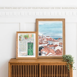 May include: Two framed watercolor paintings on a wooden shelf. The painting on the left shows a green door with a tiled wall. The painting on the right shows a cityscape with red rooftops and a blue sky.