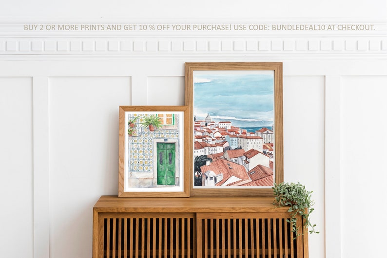 May include: Two framed watercolor paintings on a wooden shelf. The painting on the left shows a green door with a tiled wall. The painting on the right shows a cityscape with red rooftops and a blue sky.