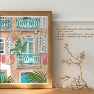 May include: Watercolour painting of a building with a balcony and colourful laundry hanging from the railing. The building has a tiled facade and green plants growing on the balcony. The painting is framed in a light wood frame.