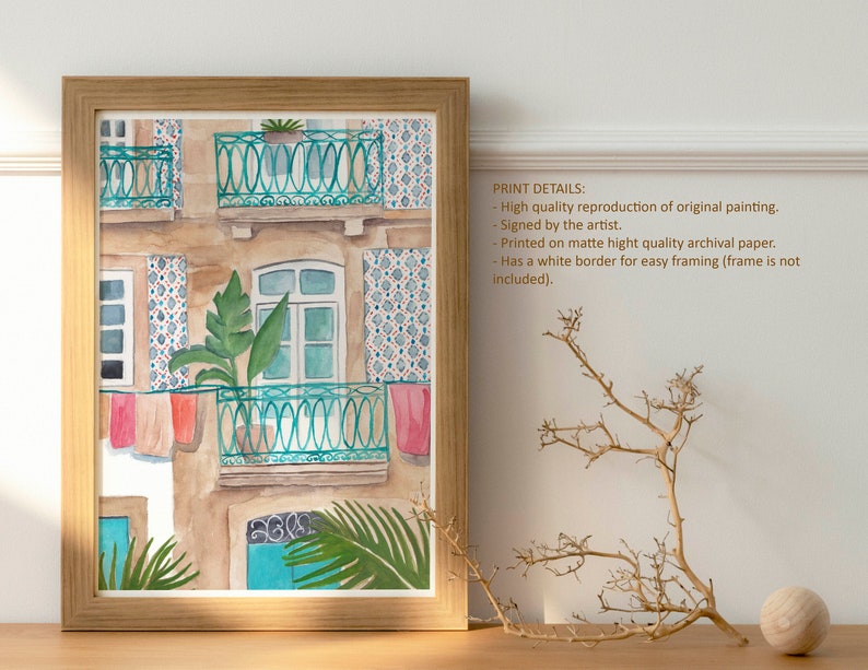 May include: Watercolour painting of a building with a balcony and colourful laundry hanging from the railing. The building has a tiled facade and green plants growing on the balcony. The painting is framed in a light wood frame.