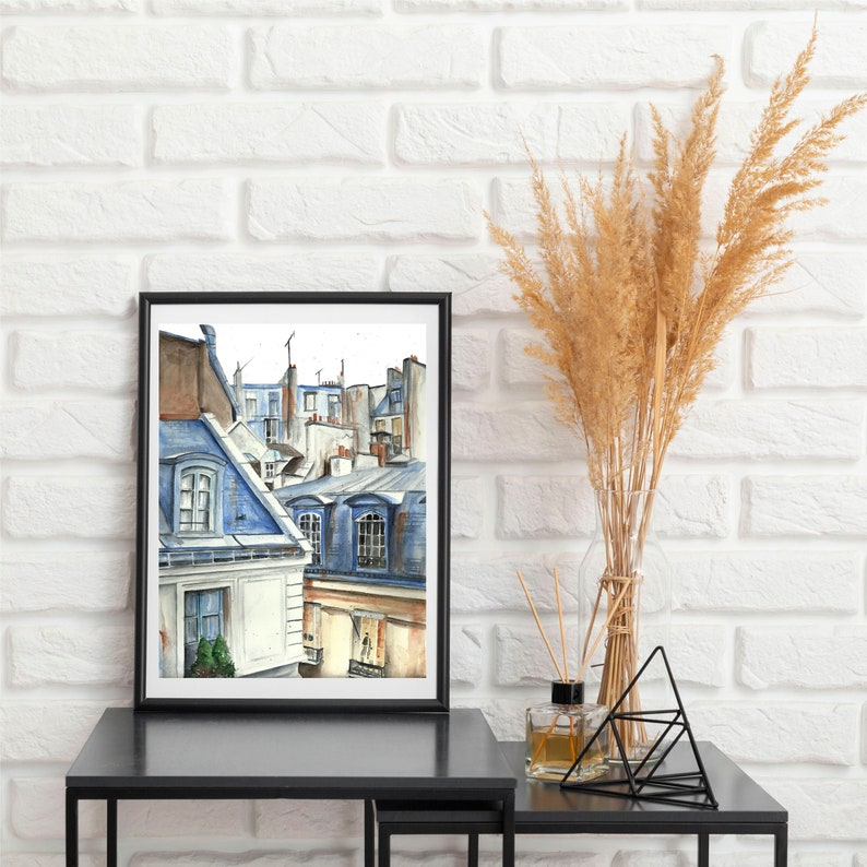 Puede incluir: Una pintura de acuarela enmarcada de un paisaje urbano parisino. La pintura representa una vista de tejados y edificios en tonos azules y grises. El marco es negro y la pintura est&aacute; colgada en una pared de ladrillo blanco.
