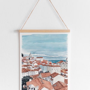 May include: A watercolor painting of a cityscape with red-tiled roofs and a blue sky. The painting is framed and hanging on a white wall.