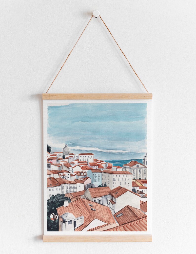 May include: A watercolor painting of a cityscape with red-tiled roofs and a blue sky. The painting is framed and hanging on a white wall.