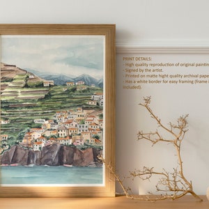 May include: A watercolor painting of a coastal town with white buildings and green hills. The painting is framed in a light wood frame and is displayed on a wooden table with a dried branch and a wooden ball.