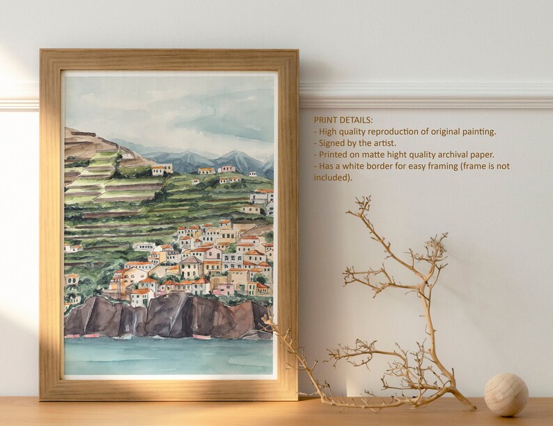 May include: A watercolor painting of a coastal town with white buildings and green hills. The painting is framed in a light wood frame and is displayed on a wooden table with a dried branch and a wooden ball.