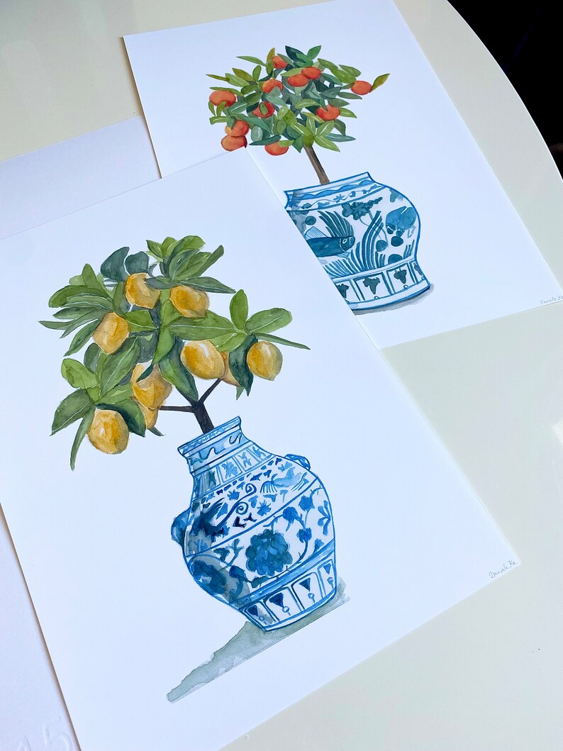 May include: Two watercolour paintings of potted citrus trees. The first painting features a small orange tree with orange fruit in a blue and white porcelain vase. The second painting features a lemon tree with yellow lemons in a blue and white porcelain vase.
