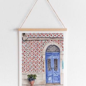 Puede incluir: Una pintura de acuarela de una puerta azul con intrincados trabajos en hierro, sobre un fondo de azulejos con patrones rojos y blancos. Una planta en maceta se encuentra a la izquierda de la puerta.