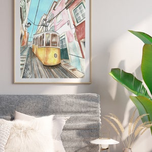May include: A watercolor painting of a yellow tram traveling up a steep, cobblestone street in Lisbon, Portugal. The buildings on either side of the street are painted in shades of pink, white, and red.