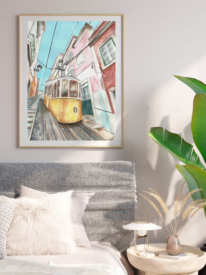 May include: A watercolor painting of a yellow tram traveling up a steep, cobblestone street in Lisbon, Portugal. The buildings on either side of the street are painted in shades of pink, white, and red.