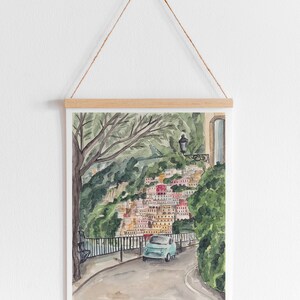 May include: A watercolour painting of a blue car driving on a winding road through a colourful village nestled in a hillside. Lush green trees and foliage surround the road and village.
