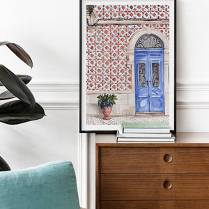 Puede incluir: Una pintura de acuarela enmarcada de una puerta azul con intrincados trabajos en hierro. La puerta est&aacute; situada contra una pared cubierta de azulejos con patrones rojos y blancos. Una planta en maceta se encuentra en el suelo frente a la puerta.