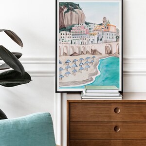 May include: A watercolor painting of a coastal town with colorful buildings, a stone archway, and a beach with blue and white striped umbrellas. The painting is framed in black and hanging on a white wall.