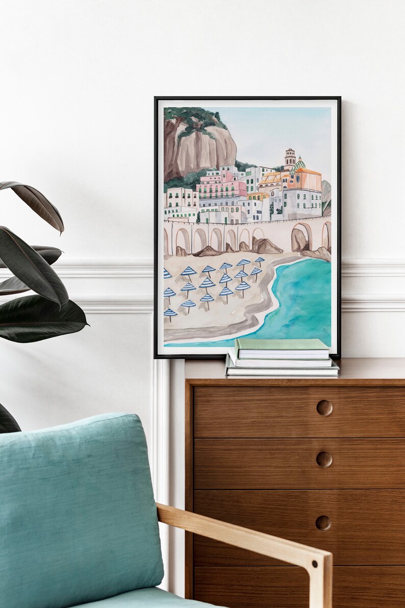 May include: A watercolor painting of a coastal town with colorful buildings, a stone archway, and a beach with blue and white striped umbrellas. The painting is framed in black and hanging on a white wall.