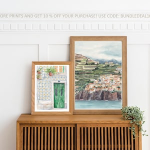 May include: Two framed watercolor paintings on a wooden shelf. The painting on the left shows a green door with a tiled wall and potted plants. The painting on the right shows a coastal town with a hillside and ocean.