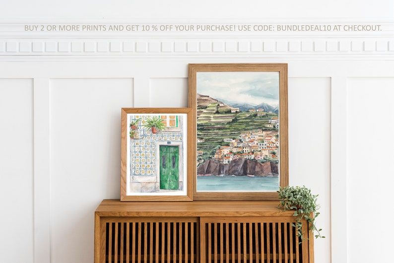 May include: Two framed watercolor paintings on a wooden shelf. The painting on the left shows a green door with a tiled wall and potted plants. The painting on the right shows a coastal town with a hillside and ocean.