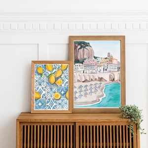 May include: Two framed watercolor paintings on a wooden shelf. The painting on the left features lemons and blue and white tiles. The painting on the right shows a colorful Italian town with a beach and turquoise water.