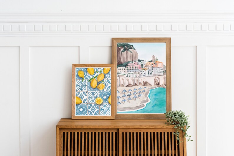 May include: Two framed watercolor paintings on a wooden shelf. The painting on the left features lemons and blue and white tiles. The painting on the right shows a colorful Italian town with a beach and turquoise water.