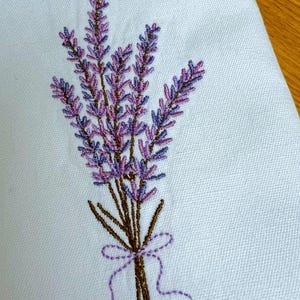 May include: Embroidered lavender bouquet in shades of purple and brown, tied with a purple ribbon. The floral design is stitched on a light blue fabric, showcasing a detailed botanical pattern. The embroidery adds a touch of elegance.