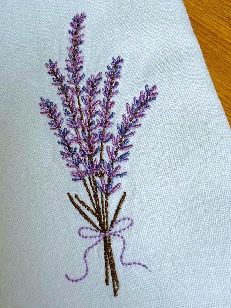 May include: Embroidered lavender bouquet in shades of purple and brown, tied with a purple ribbon. The floral design is stitched on a light blue fabric, showcasing a detailed botanical pattern. The embroidery adds a touch of elegance.