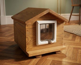 Puede incluir: Una pequeña caseta de madera para mascotas con techo a dos aguas y una puerta de plástico blanco instalada en el panel frontal. La estructura es de madera marrón claro con veta visible y se asienta sobre una base, con una solapa cuadrada transparente.
