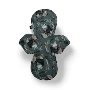 May include: A green reusable cloth menstrual pad with a whimsical frog and floral print. The pad has a snap closure and is made of soft, absorbent fabric.