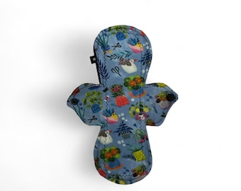 Puede incluir: Una compresa menstrual de tela reutilizable azul con estampado floral y de pájaros. La compresa tiene un ribete negro y broches a los lados.