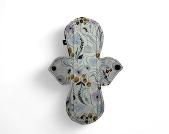 Puede incluir: Una compresa menstrual de tela reutilizable blanca con un patrón floral en tonos de morado, marrón y amarillo. La compresa tiene broches negros a los lados y una etiqueta negra en la parte inferior.