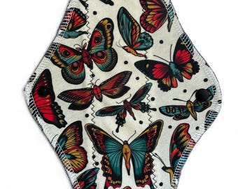 May include: A white reusable cloth pad with a colorful butterfly print. The butterflies are in shades of red, blue, yellow, and green. The pad has a black stitched edge.