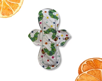 May include: A reusable cloth menstrual pad featuring a white fabric base with a repeating pattern of green caterpillars and colorful polka dots. The pad has a contoured shape with side wings and is stitched with a dark grey thread along the edges.