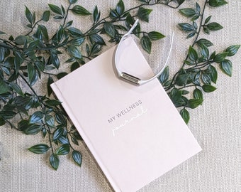 May include: A pink wellness journal with the text "My Wellness Journal" printed on the cover. A silver fitness tracker is lying on top of the journal.