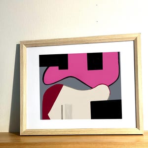 May include: Abstract art print in a light wood frame. The artwork features geometric shapes in pink, black, white, grey, and burgundy. The framed art is displayed against a white wall.