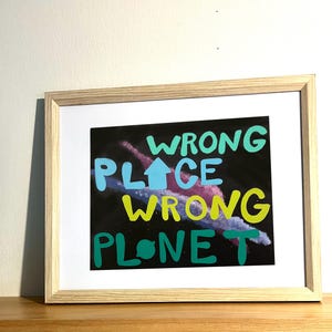 Puede incluir: Impresión artística enmarcada con las palabras "WRONG PLACE WRONG PLANET" en verde azulado, amarillo y verde. La obra de arte presenta un diseño abstracto y colorido sobre un fondo negro. La impresión está en un marco de madera clara.