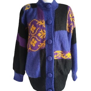 Könnte beinhalten: Vintage Cardigan mit Knopfleiste im Patchwork-Design. Der Pullover hat einen hohen Kragen und große Knöpfe. Das Farbschema umfasst Schwarz, Lila und Blau mit goldenen Akzenten. Der Cardigan wird auf einer Schaufensterpuppe präsentiert.